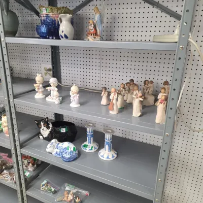 Sale Photo Thumbnail #23: Collection of collectible figurines and ceramics displayed on grey metal shelving, featuring Willow Tree carved-look angels, Precious Moments porcelain figures, and decorative cat planters.