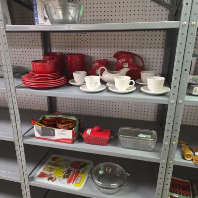 Sale Photo Thumbnail #38: Collection of kitchenware displayed on metal shelving, featuring red Fiesta-style dishware, white teacups with saucers, glassware, and decorative trays.