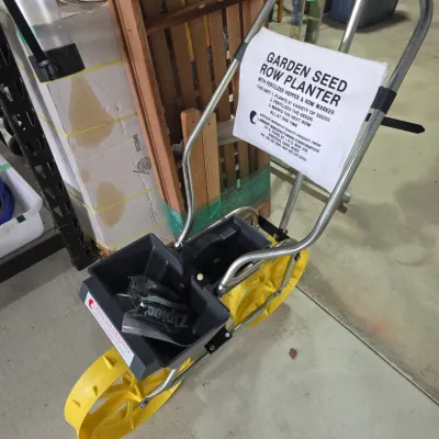 Sale Photo Thumbnail #112: Walk-behind garden seed row planter featuring yellow plastic wheels, double hoppers, tubular metal handle frame, and attached informational sheet.