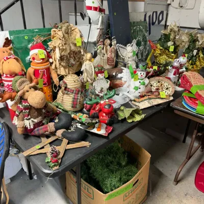 Sale Photo Thumbnail #29: Collection of rustic and folk-style Christmas decorations, including plush snowmen, reindeer figurines, angels, and faux garland displayed on folding tables.