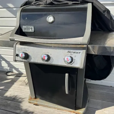 Sale Photo Thumbnail #34: Weber Spirit three-burner propane gas grill with side shelves, integrated lid thermometer, and a black protective cover.