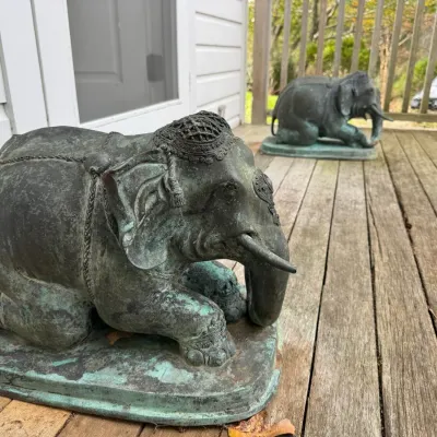 Sale Photo Thumbnail #8: Pair of large weathered bronze kneeling elephant statues with decorative caparisons and a heavy verdigris patina, displayed outdoors on a wooden deck.