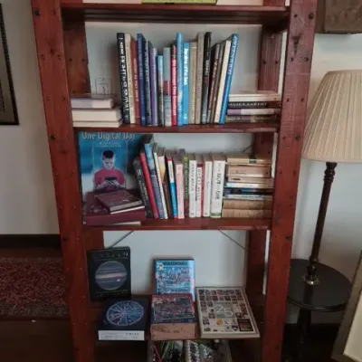 Sale Photo Thumbnail #103: Tall reddish-brown wooden bookshelf holding various hardcover and paperback books, boxed games, and art supplies.