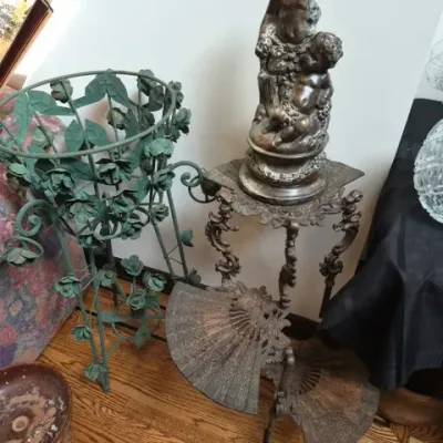 Sale Photo Thumbnail #120: Vintage classical cherub statue displayed on a ornate cast metal pedestal stand beside a green wrought-iron plant stand.