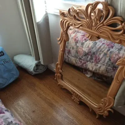 Sale Photo Thumbnail #80: Ornately carved light wood wall mirror in a Baroque style resting on hardwood flooring next to a blue AeroBed storage bag.