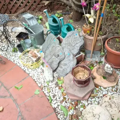 Sale Photo Thumbnail #188: Collection of outdoor garden decor including concrete stepping stones, plastic watering cans, potted plants, and wrought iron plant stands.