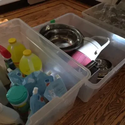 Sale Photo Thumbnail #135: Clear plastic storage totes packed with household goods, including cleaning supplies, metal mixing bowls, a insulated beverage pitcher, and glassware.