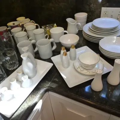 Sale Photo Thumbnail #128: Large collection of white ceramic dinnerware, coffee mugs, salt and pepper shakers, and clear glassware set out on a countertop.