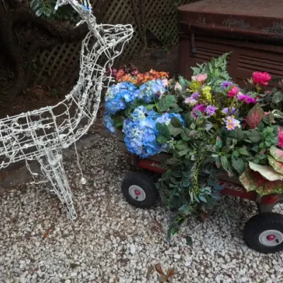 Sale Photo Thumbnail #186: Wireframe lighted reindeer Christmas decoration standing next to a red Radio Flyer wagon filled with artificial flowers and greenery.