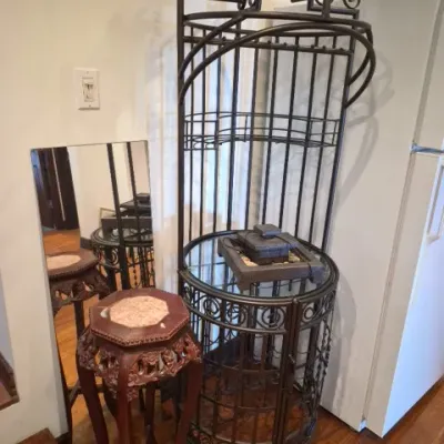Sale Photo Thumbnail #57: Tall wrought iron corner baker's rack featuring decorative scrollwork, alongside an carved wooden plant stand with an inlaid marble top.