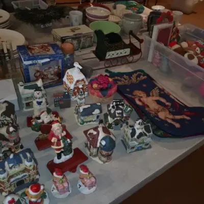 Sale Photo Thumbnail #160: A table full of holiday decorations including Christmas village buildings, Santa figurines, an embroidered stocking, dishware, and plush toys.