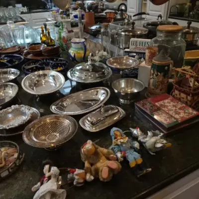 Sale Photo Thumbnail #124: Large collection of silver-plated serving dishes, bowls, and trays displayed on a countertop alongside vintage collectibles, Wizard of Oz dolls, and glassware.