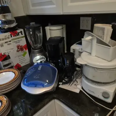 Sale Photo Thumbnail #132: Collection of kitchen counter appliances including Cuisinart and Oster blenders, a coffee maker, a Black & Decker steamer, and stacked patterned dinnerware.