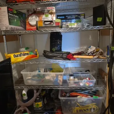 Sale Photo Thumbnail #189: Four-tier wire shelving unit filled with household items, including an APC battery backup, First Alert smoke alarms, Duraflame log, and plastic storage tubs with assorted supplies.