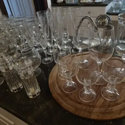 Sale Photo Thumbnail #129: Large collection of glassware including champagne flutes, wine glasses, and brandy snifters displayed on a countertop next to a wooden tray and crystal shakers.