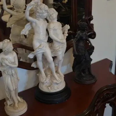 Sale Photo Thumbnail #116: Collection of classical-style statues, including a large white figural group of dancing cherubs, a smaller white female classical maiden figure, and a dark bronze-toned female figure, displayed on a wooden console table.