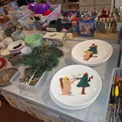 Sale Photo Thumbnail #159: Assorted household goods, including whimsical illustration plates, glassware, a peacock feather fan, and hand tools arranged on a table for sale.