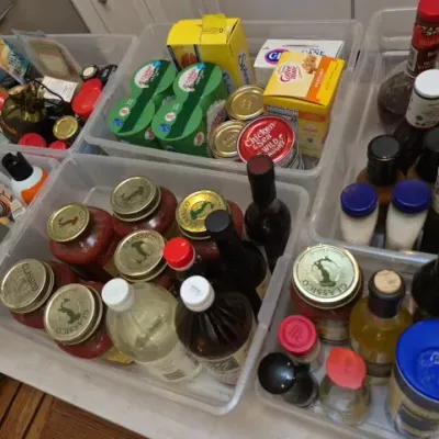 Sale Photo Thumbnail #134: Clear plastic storage bins filled with assorted pantry goods, including bottled condiments, canned seafood, pasta sauces, and sweeteners.