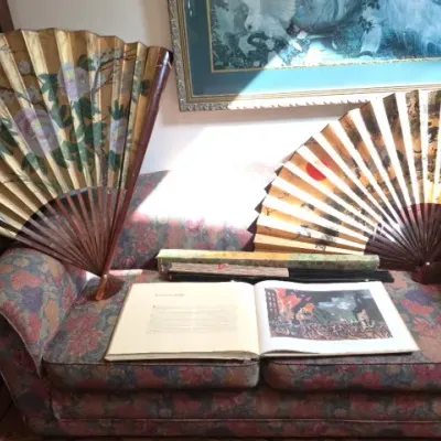 Sale Photo Thumbnail #118: Pair of large Asian decorative hand fans with painted floral and landscape motifs on gilt paper, displayed on a patterned sofa alongside an open art history book and folded fans.