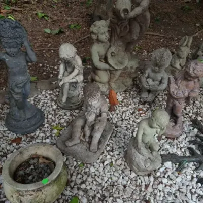 Sale Photo Thumbnail #187: Collection of concrete and stone garden statues, featuring cherubs, children, and classic figures arranged in a outdoor gravel bed.