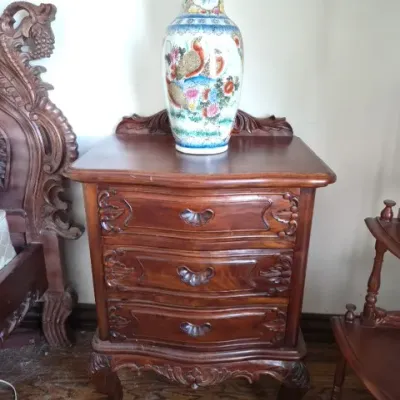 Sale Photo Thumbnail #34: Ornately carved wooden three-drawer nightstand in a dark finish, topped with a tall Chinese porcelain baluster vase featuring hand-painted floral and bird motifs.