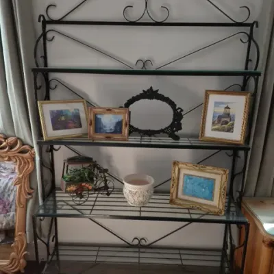 Sale Photo Thumbnail #107: Black wrought iron baker's rack with scrollwork detailing and glass shelves, featuring framed artwork and decorative home accents.
