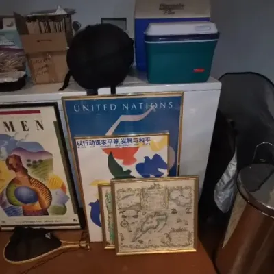 Sale Photo Thumbnail #157: Collection of framed art prints including United Nations themed posters, alongside coolers, household items, and a stainless steel trash can.