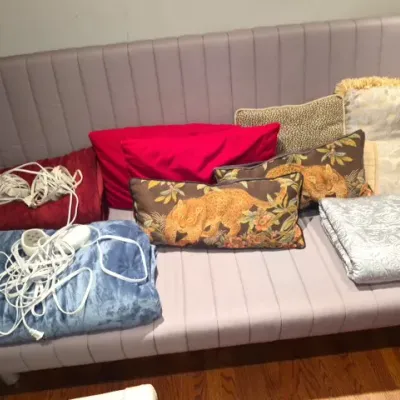 Sale Photo Thumbnail #142: A modern channel-tufted mauve armless sofa lined with an assortment of throw pillows, electric heated blankets, and folded linens.