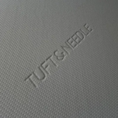 Sale Photo Thumbnail #111: Close-up of a Tuft & Needle mattress cover featuring embossed brand lettering on textured light grey fabric.