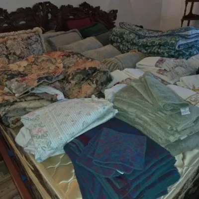 Sale Photo Thumbnail #141: An assortment of household linens, folded quilts, floral throw pillows, and bath towels laid out on a dark carved wooden bed frame.