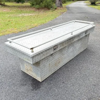 Aluminum diamond-plate crossover truck tool box featuring an attached top cargo rail and side latches, showing minor weathering and...