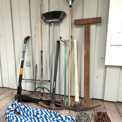 Collection of long-handled lawn and yard tools, including rakes, brooms, pickaxes, along with coiled blue and white rope and metal chains.