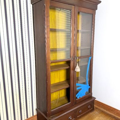 Vintage dark wood display cabinet featuring glass doors, a yellow interior back, and bottom storage drawers.