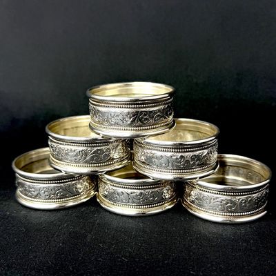 Set of six vintage silver-plated napkin rings featuring ornate embossed floral and scrollwork detailing with beaded borders.