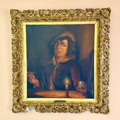 Framed oil painting in the Dutch Golden Age genre style, depicting a figure seated at a table illuminated by candlelight. Housed in an...