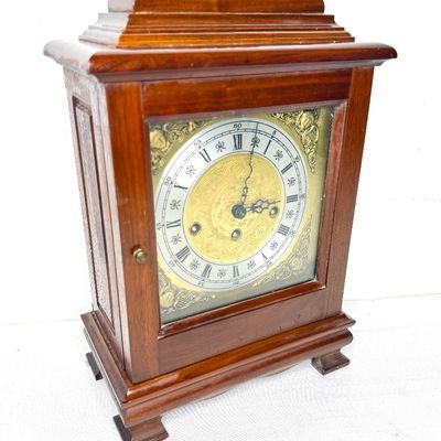 Traditional wooden mantle clock featuring an ornate brass dial with Roman numerals, a decorative top handle, and a polished wood case.
