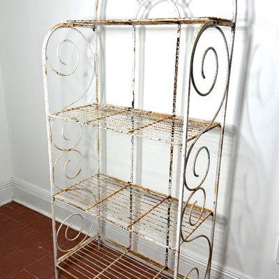 Vintage white wrought iron baker's rack featuring four wire shelves, scrollwork side accents, and an arched sunburst crest, showing...