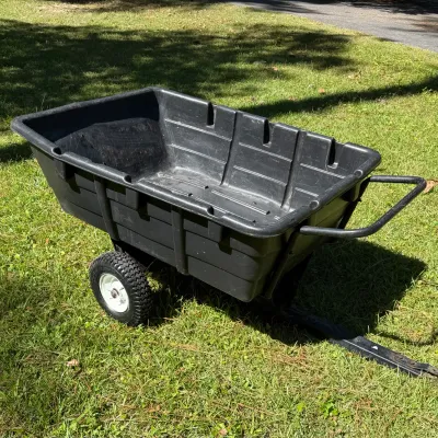 Heavy-duty black plastic utility dump cart with pneumatic tires and a tow hitch attachment, showing light wear from use.