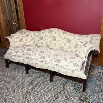 Chippendale-style Camelback sofa upholstered in cream floral fabric, featuring carved mahogany legs, nailhead trim, and matching throw...