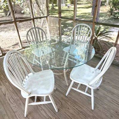 Glass top patio dining set featuring a round glass-top table with a distressed white metal base and four distressed white wooden Windsor...