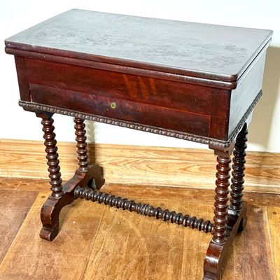 Antique mahogany flip-top game or sewing table featuring spool-turned legs and stretcher, with visible surface wear to the finish.