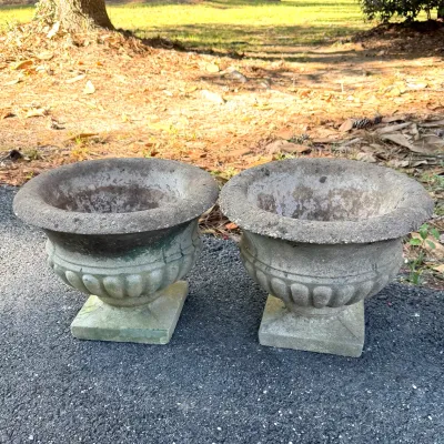 Pair of classic cast concrete urn planters with fluted bodies and square pedestal bases, showing weathered patina and moss accents.