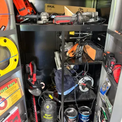 Sale Photo Thumbnail #68: Black metal storage cabinet filled with assorted power tools, lawn care equipment, and paint cans, including a hedge trimmer, circular saw, angle grinder, and garden sprayer.