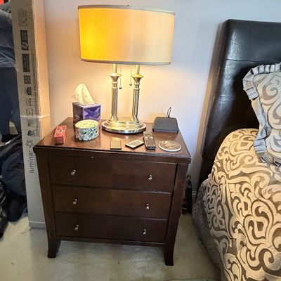 Sale Photo Thumbnail #309: Dark brown wood three-drawer nightstand with silver knobs, topped with a brushed metal table lamp featuring a oval drum shade.