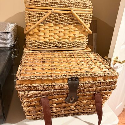 Sale Photo Thumbnail #236: Stack of two woven wicker picnic hampers featuring wooden handles, fabric shoulder straps, and a front latch closure.