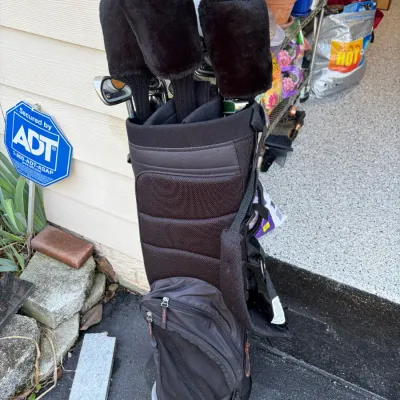 Sale Photo Thumbnail #25: Black TaylorMade golf stand bag loaded with clubs, including woods with plush black headcovers.