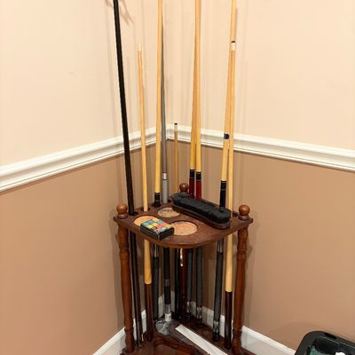 Sale Photo Thumbnail #245: Corner wooden pool cue rack holding multiple billiard sticks, a bridge stick, felt brush, and chalk.