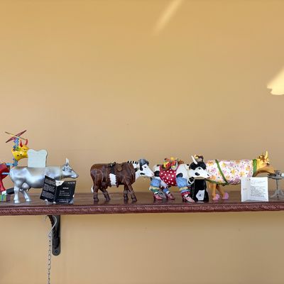 Sale Photo Thumbnail #124: Collection of decorative CowParade resin figurines lined up on a wooden shelf, featuring various artistic themes and designs.