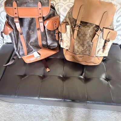 Sale Photo Thumbnail #307: Two Coach leather and canvas backpacks, including a dark brown horse and carriage print model and a tan signature C print model, resting on a tufted leather bench.