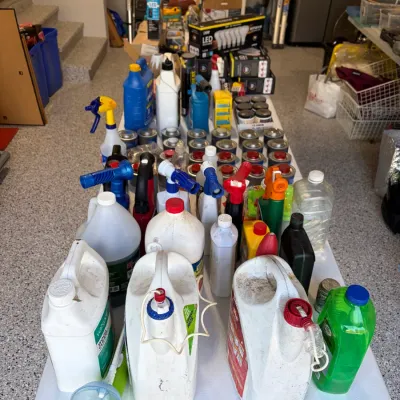 Sale Photo Thumbnail #86: Large assortment of household cleaning supplies, lawn chemicals, paint cans, and automotive fluids arranged on a white folding table in a garage.